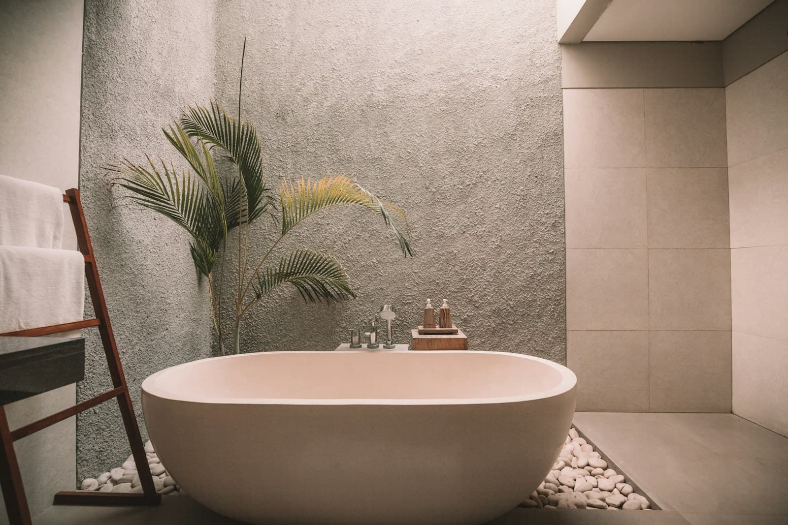 A luxury spa-style bathroom with a freestanding oval tub, stone walls, palm plant, white pebbles, and ambient candlelight