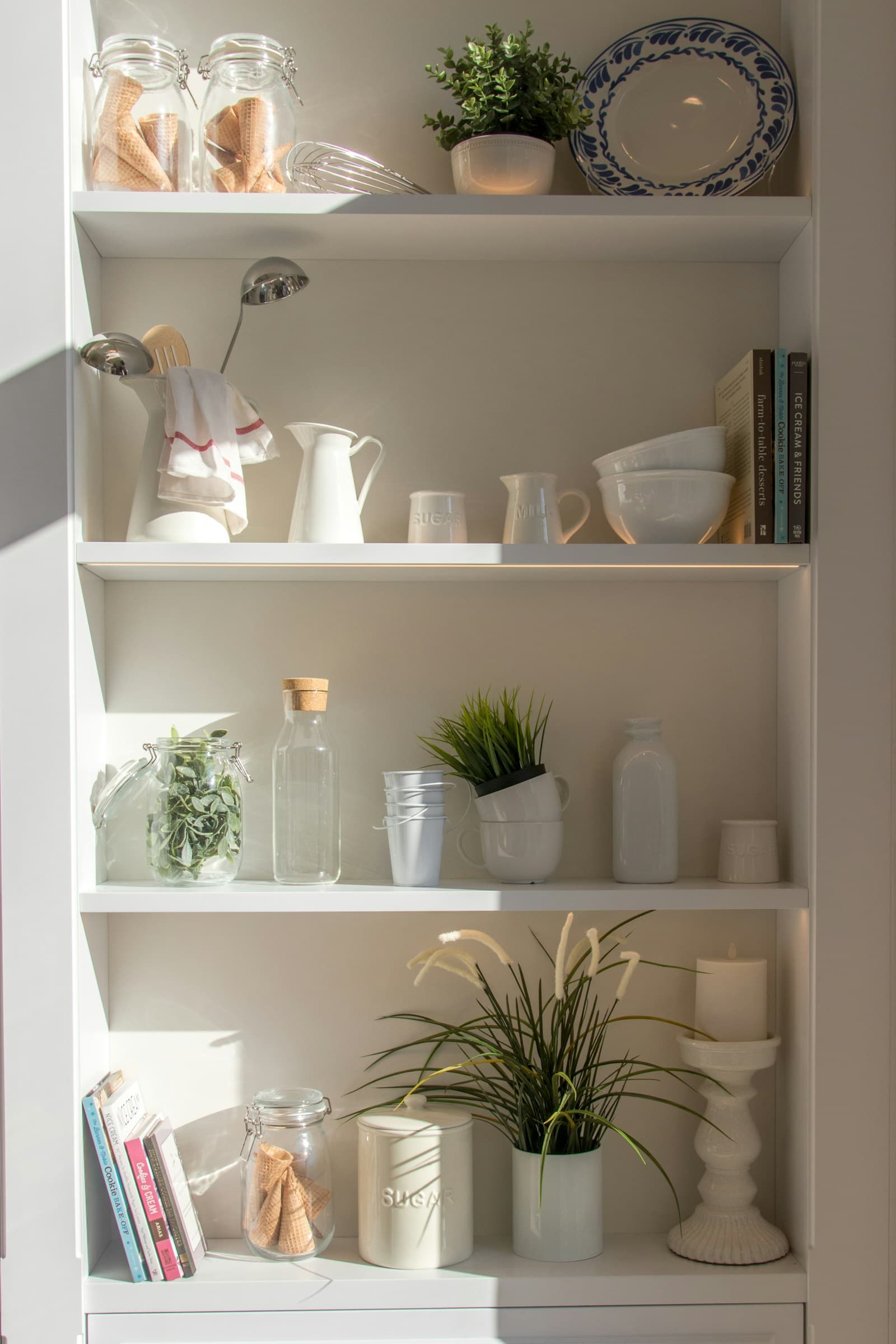 Elegant white built-in shelving with curated kitchen items, ceramics, cookbooks, and potted herbs arranged artfully